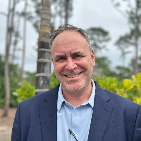 Bruno E. Pouliot smiling in a navy blazer and light-blue shirt, outdoors beside a palm trunk and greenery